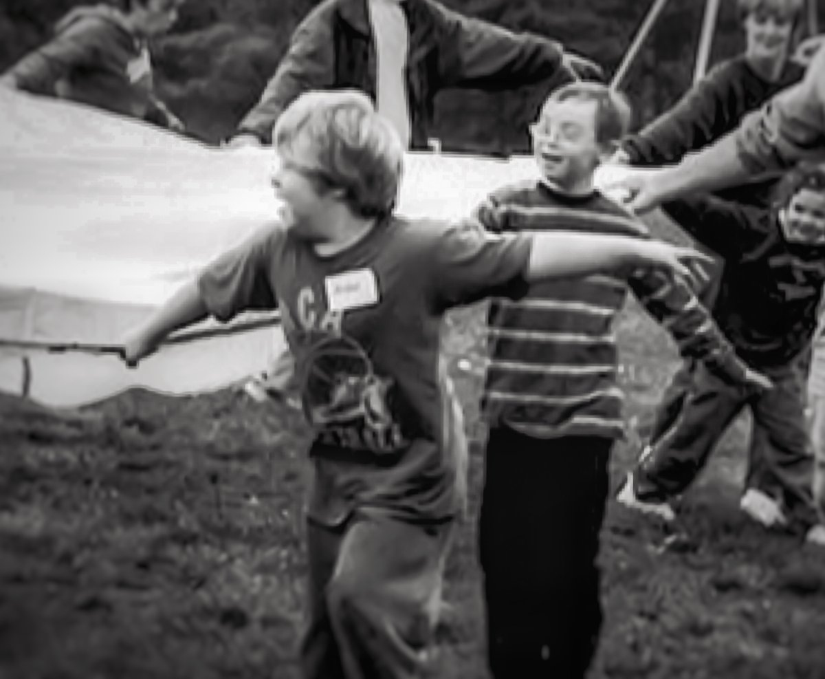 Black and white photo of a group of children cheerfully holding onto a play parachute in a field.