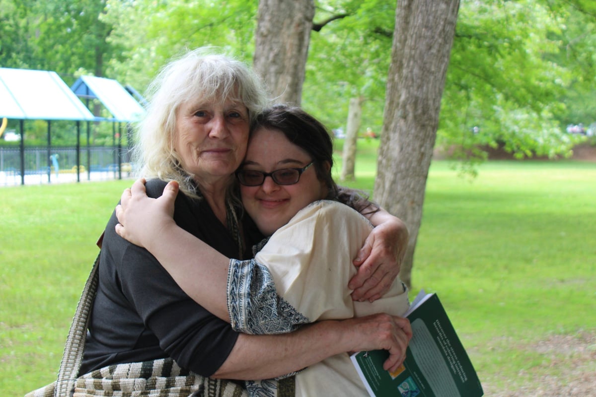 A young woman with a disability, a light skin tone and shoulder-length brown hair in a braid, smiles as she hugs an smiling older woman with light skin tone and white, chin-length hair.