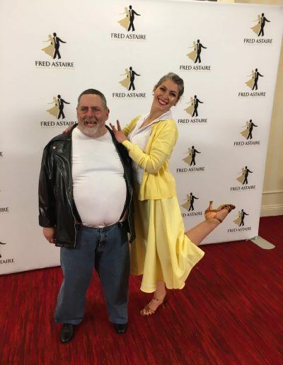 Michael Cooley, an older man with a disability, light skin tone, and short gray hair, smiles while he poses on a red carpet with a woman with a light skin tone and short, blond hair. She is smiling and kicking up one of her legs as she holds Michael’s shoulders.