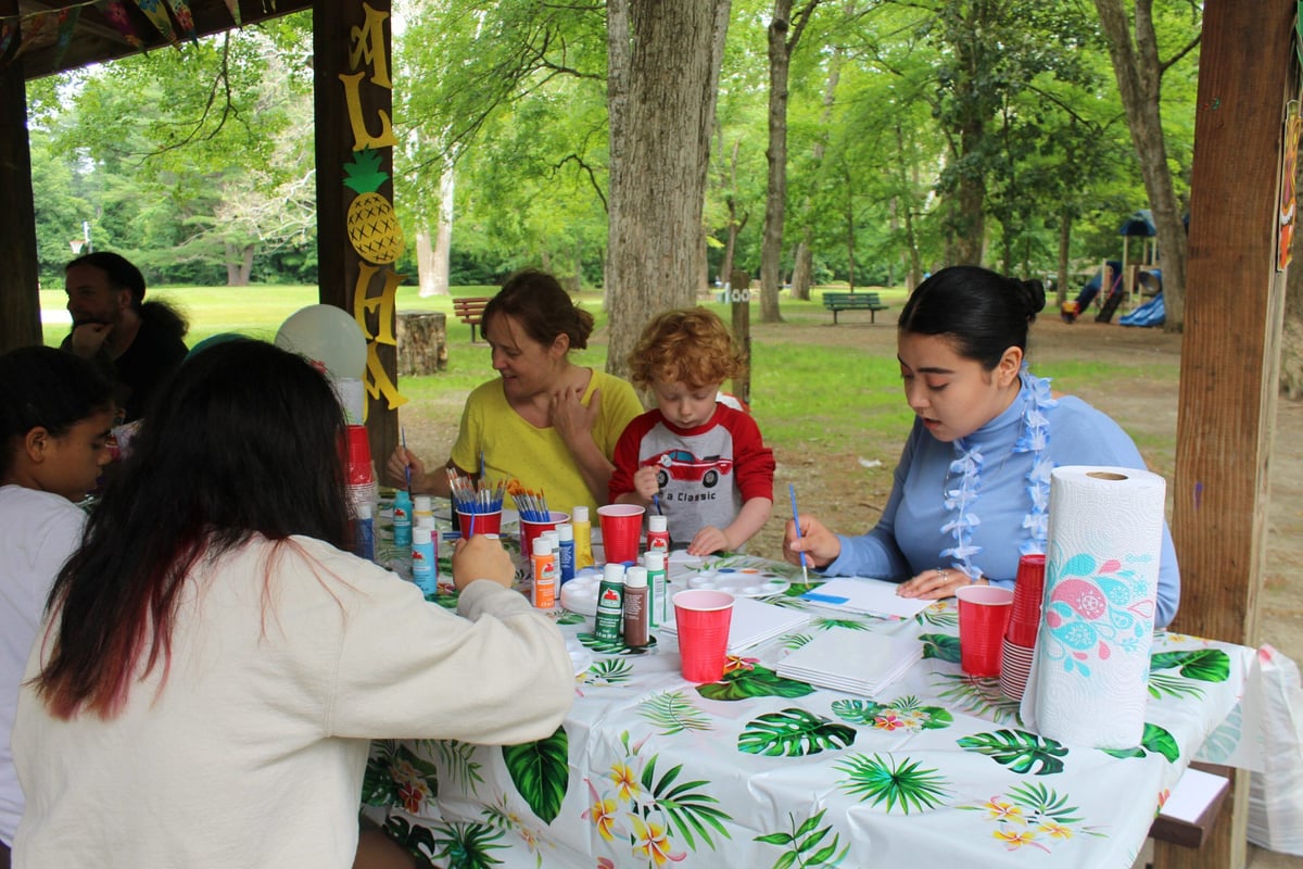 A diverse group of children sit at a picnic table in a park, painting.