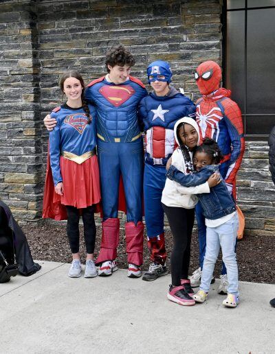 Four people dressed up as Wonder Woman, Superman, Captain America and Spider man stand with their arms around each other’s shoulders. In front of them are tow young girls with dark skin tones and black hair, hugging each other.