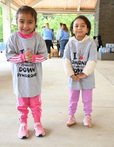 Two young girls with medium skin tone and short brown smile toward the camera while standing underneath an open building in a park.