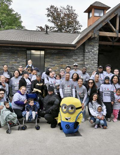 A diverse group of people stand in front of a cabin. In the front of the group is a minion sitting on the ground.