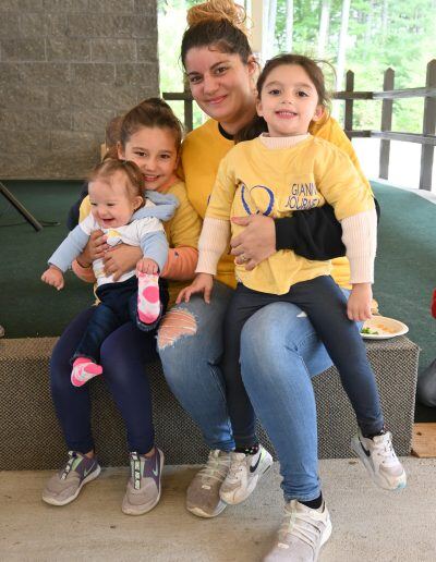 A young woman with tanned skin and brown hair in a bun holds two young girls and an infant with lighter skin tones on her lap. The woman and girls are wearing yellow shirts and smiling.