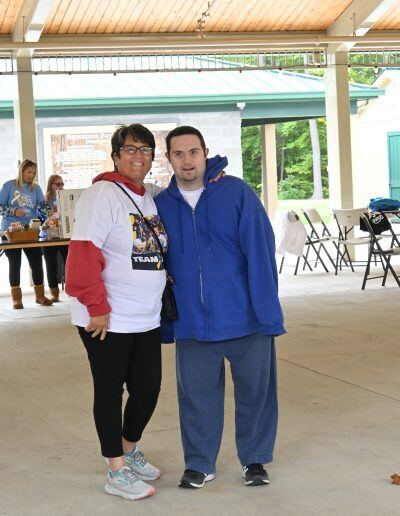A woman with tanned skin and short brown hair and a man with a light skin tone and short brown hair hold each other at the shoulders. They are smiling at the camera while standing underneath an open building in a park.
