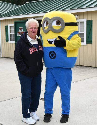 An older man with a light skin tone stands next to a minion mascot.