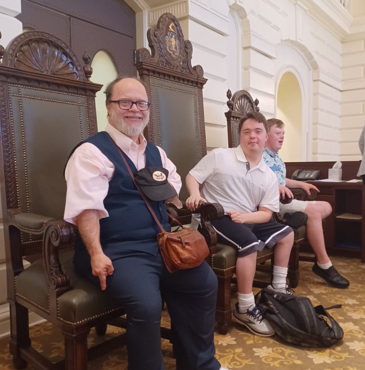 Three men of various ages, with disabilities, and light skin tones, sit in seats of honor at the Massachusetts State House.