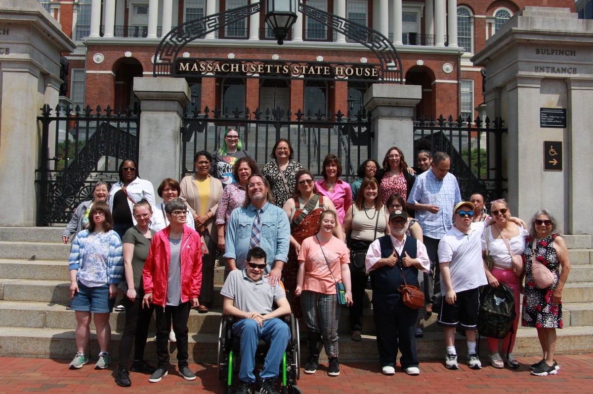 A large, diverse group stands together on the stairs of the Massachusetts State House.