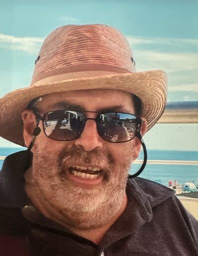 Closeup of Michael Cooley, an older man with a disability. He has a light skin tone and short gray hair and wears a straw hat with sunglasses on the beach while smiling.