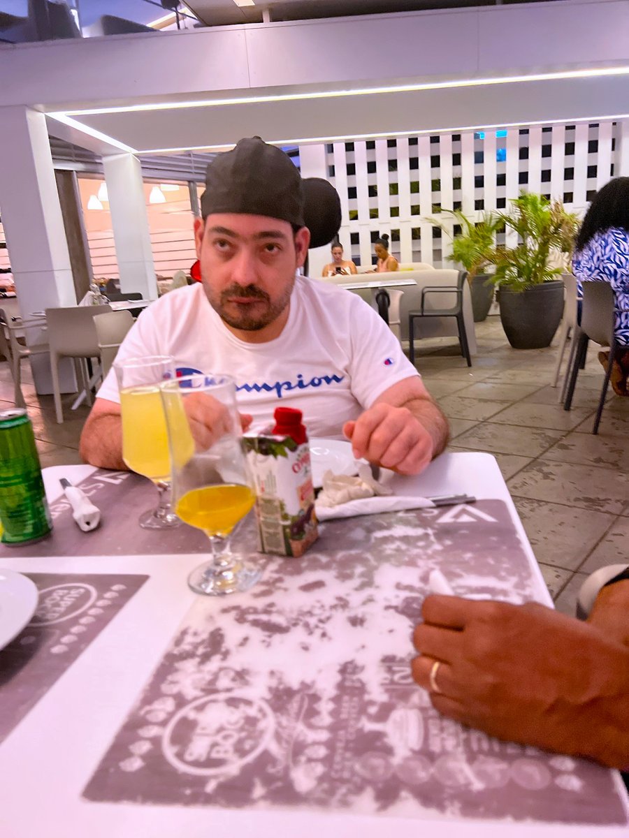 Gadriel, a man with a disability and light skin tone, sits a restaurant table. In front of him are several colorful drinks in cocktail glasses.