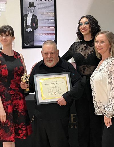 Michael Cooley, an older man with a disability, light skin tone, and short gray hair, holds a certificate while standing next to three smiling women with light skin tones and wearing various evening dresses.