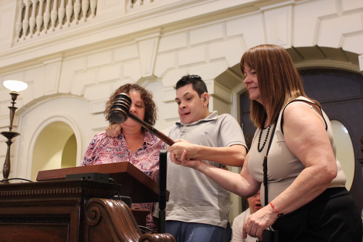 A man with a disability, light skin tone, and short brown hair swings a gavel at the Massachusetts State House. He is assisted by a woman with a light skin tone and shoulder-length brown hair.
