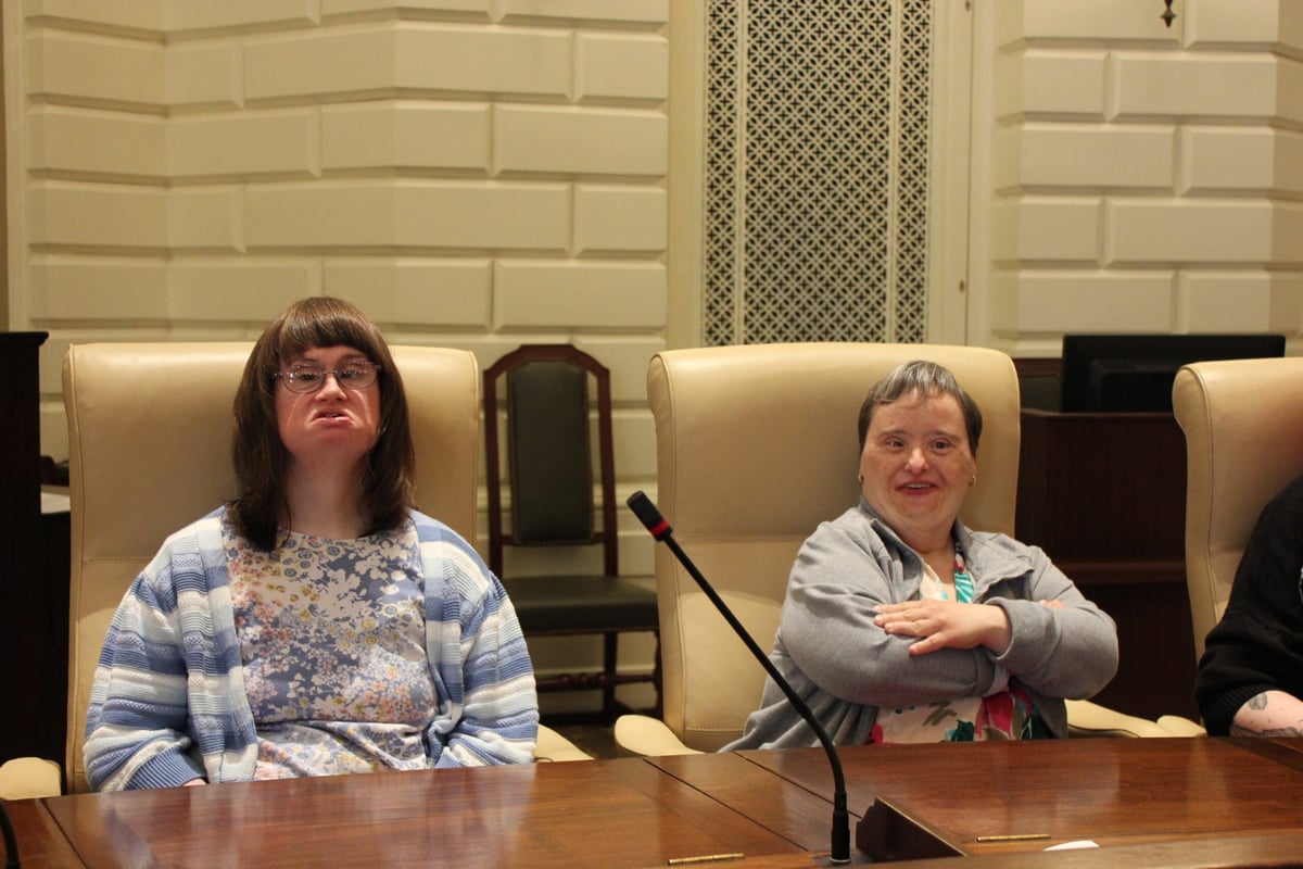 Two women with disabilities and light skin tones sit in the chairs of senators at the Massachusetts State House.