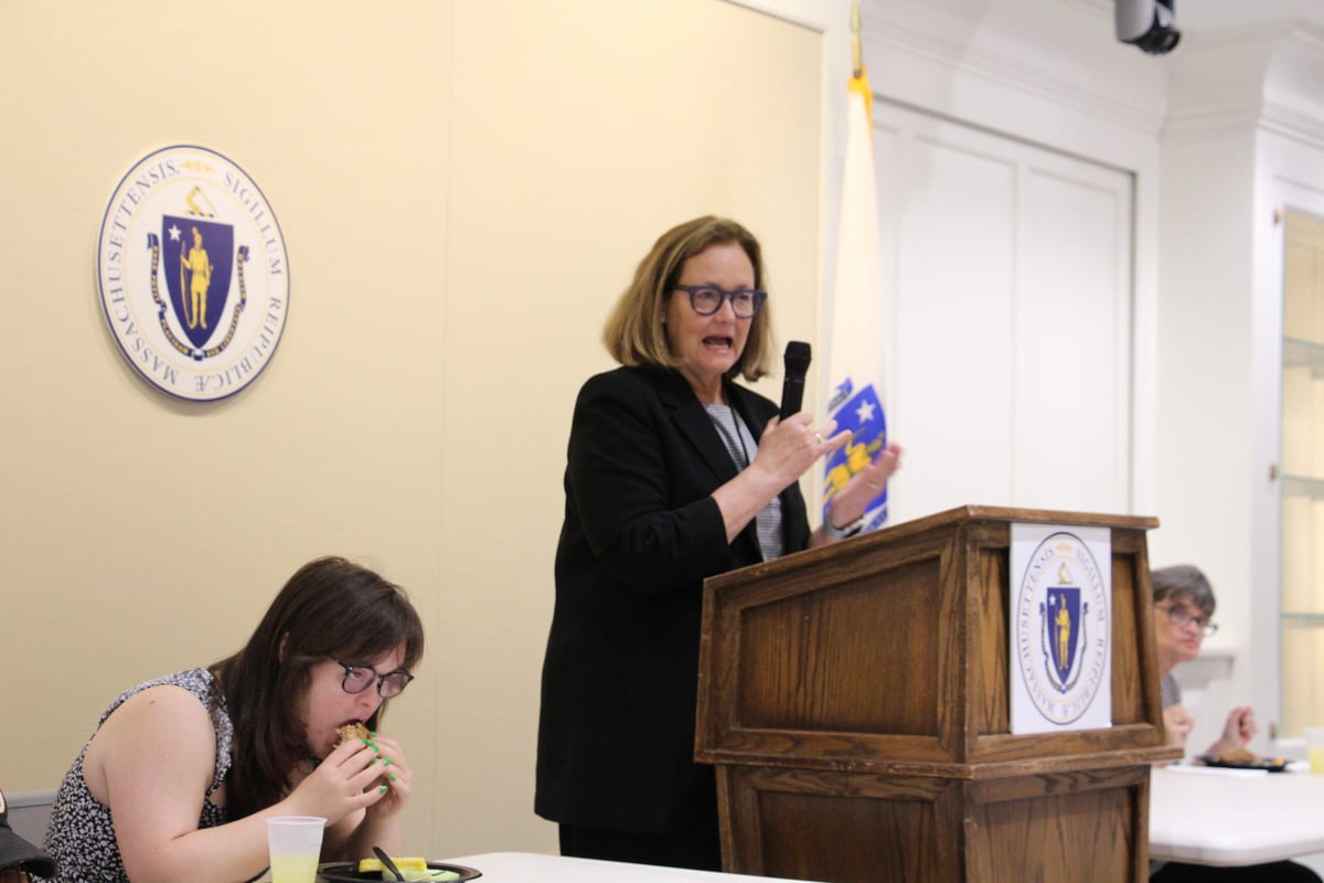 Secretary of Health and Human Services, Kate Walsh, is a middle-aged woman with a light skin tone and chin-length blond hair. She speaks at a podium at the Massachusetts State House.