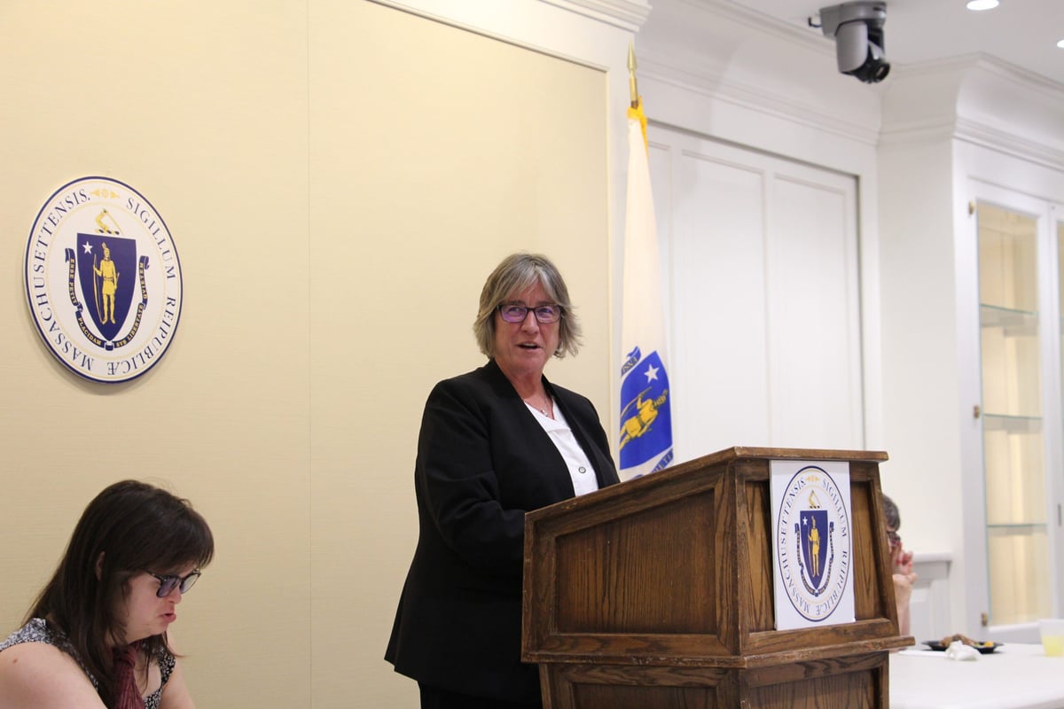 Commissioner of the Department of Development Services, Jane Ryder, is a middle-aged woman with a light skin tone and short gray hair. She speaks at a podium at the Massachusetts State House.