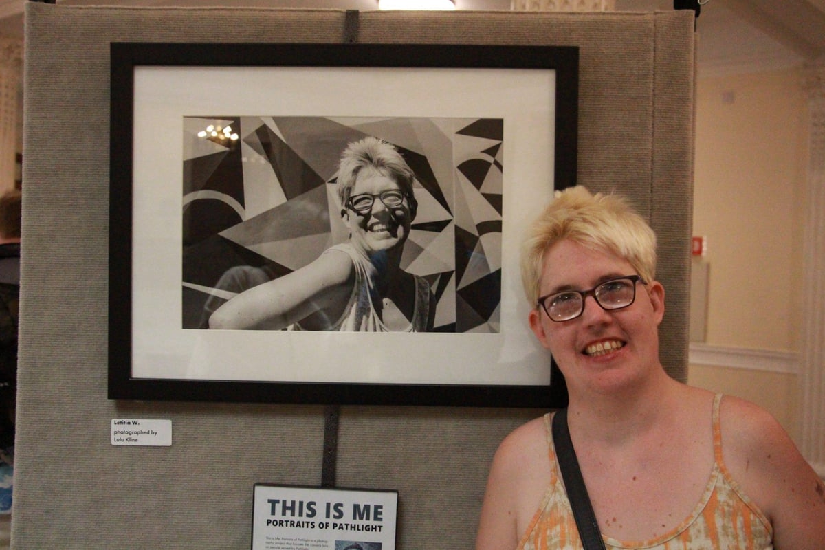 Letitia Ward, a woman with a disability, a light skin tone, and short bleached hair, stands and smiles in front of a black-and-white photo of herself.