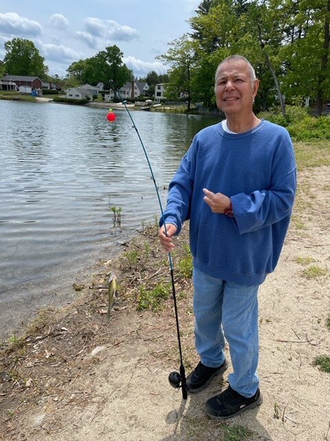 A man with a disability and a light skin tone stands in front of a lake while holding a fishing pole. He is wearing a blue sweater.