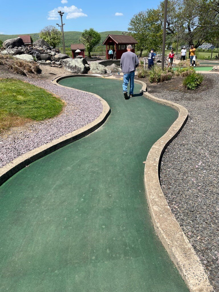 A man with a light skin tone walks toward the end of a mini golf hole.