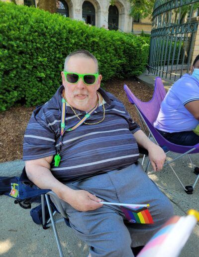 Michael Cooley, an older man with a disability, light skin tone, and short gray hair, sits in a camping chair while holding a progress flag.