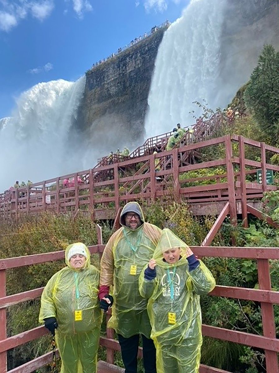 Three people with light skin tones stand in front of Niagara Falls on red walkways while wearing plastic yellow ponchos.