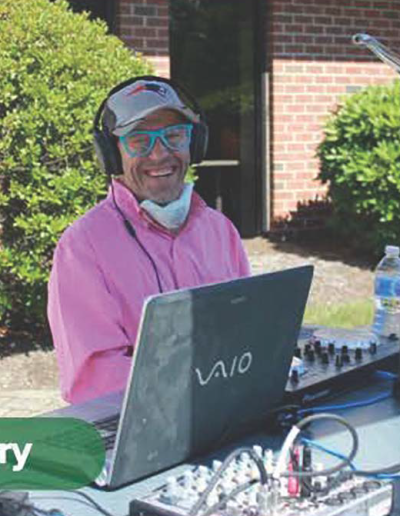 A man with a light skin tone wearing headphones and a pink shirt stands behind a laptop and DJ mixing table while smiling at the camera.