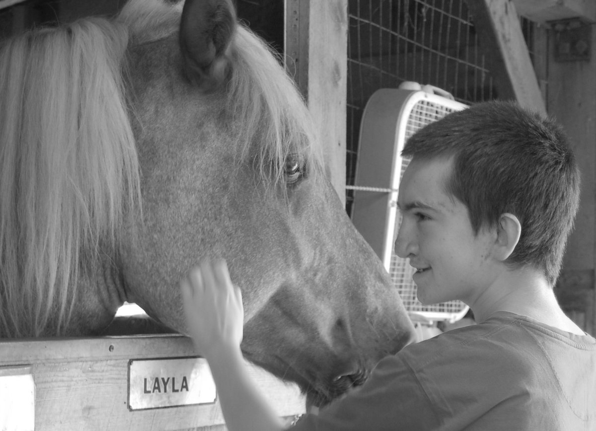 Black and white photo of a teenager with a light skin tone petting a horse’s cheek.