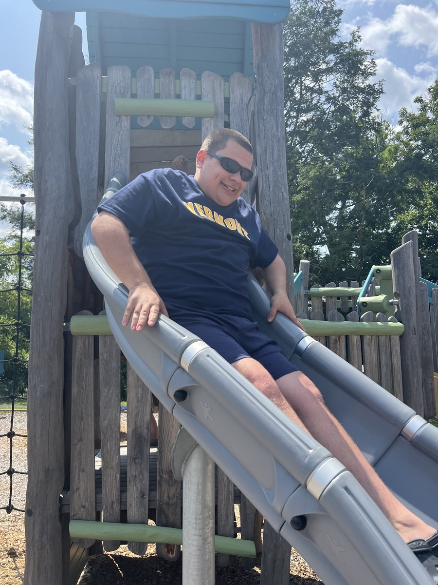 A man with a disability and a light skin tone smiles as he rides a playground slide.