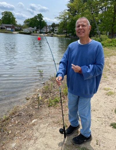 A man with a disability and a light skin tone stands in front of a lake while holding a fishing pole. He is wearing a blue sweater.