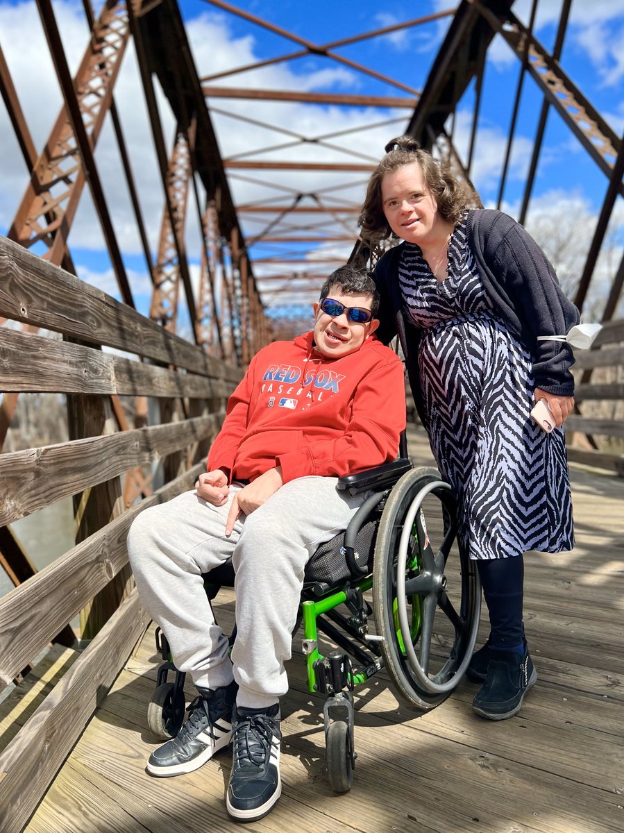 A man using a wheelchair with a light skin tone and an older woman with a light skin tone pose while standing on the Norwottuck walking bridge.