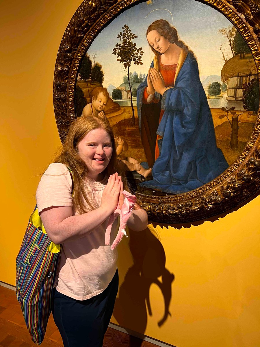 A young woman with a disability and a light skin tone mimics a hand prayer while standing in front of a painting at the Smith Art Museum.