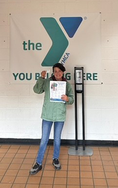 A woman with a disability and a light skin tone holds a lanyard and pamphlet for a YMCA membership in front of a YMCA banner.