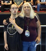 A man with a disability and a light skin tone stands in front of a stage before a rock concert. An older woman with a light skin tone and long, white hair stands beside him, keeping an arm around his shoulder.