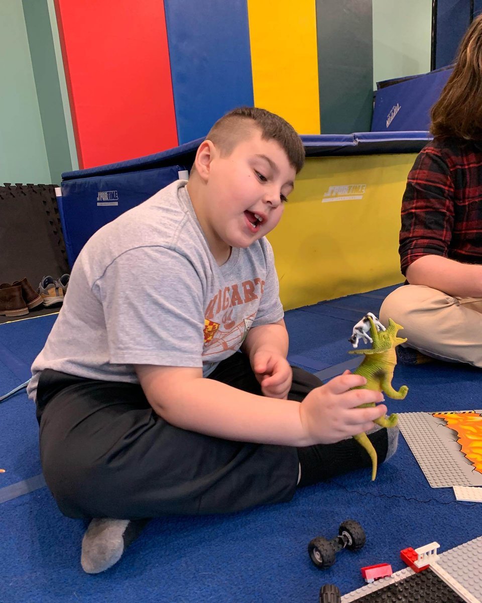 A boy with a light skin tone sits with crossed legs and plays with a triceratops toy dinosaur.