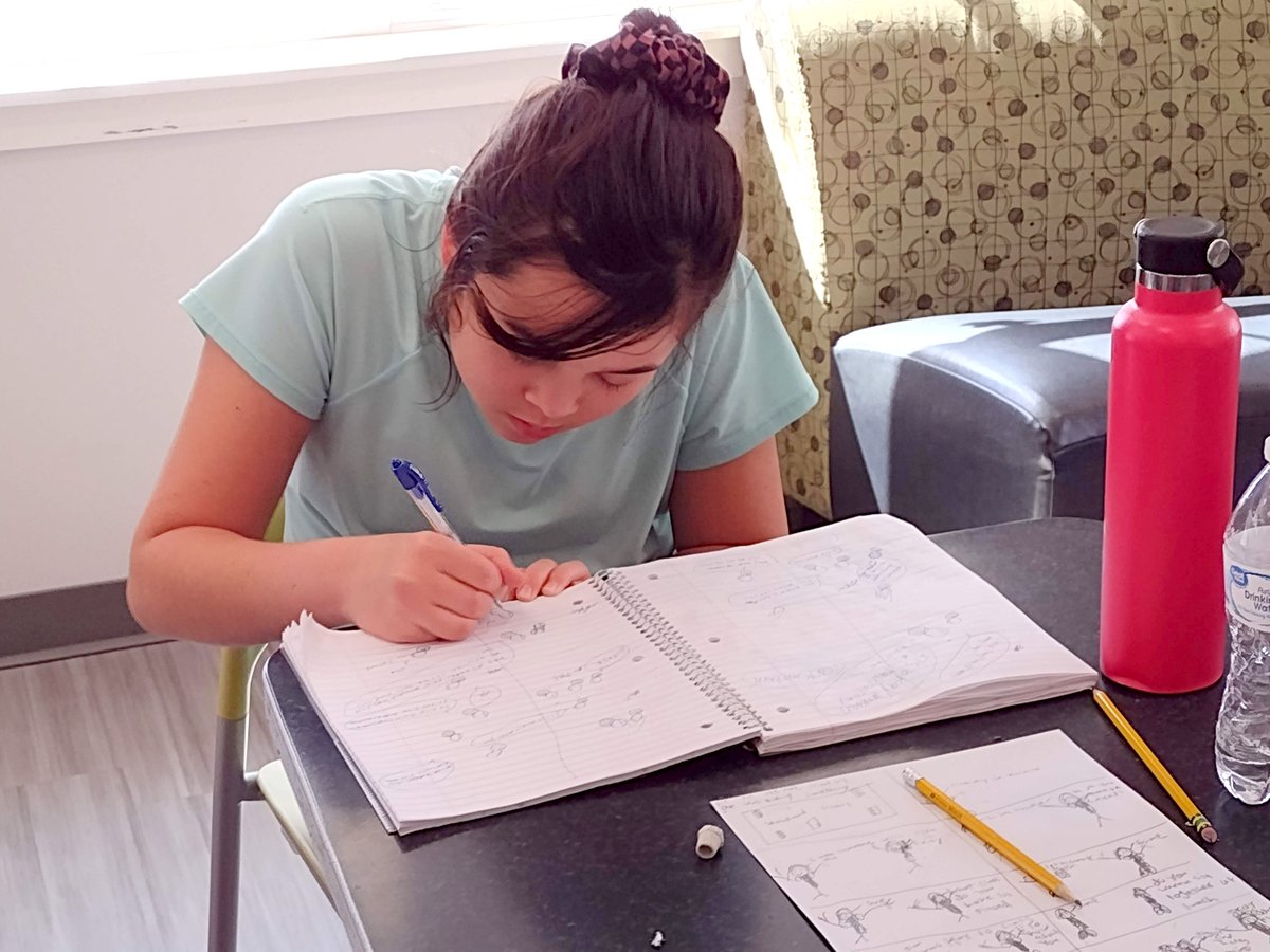 A woman with a light skin tone focuses on writing in a notebook while sitting in a chair.