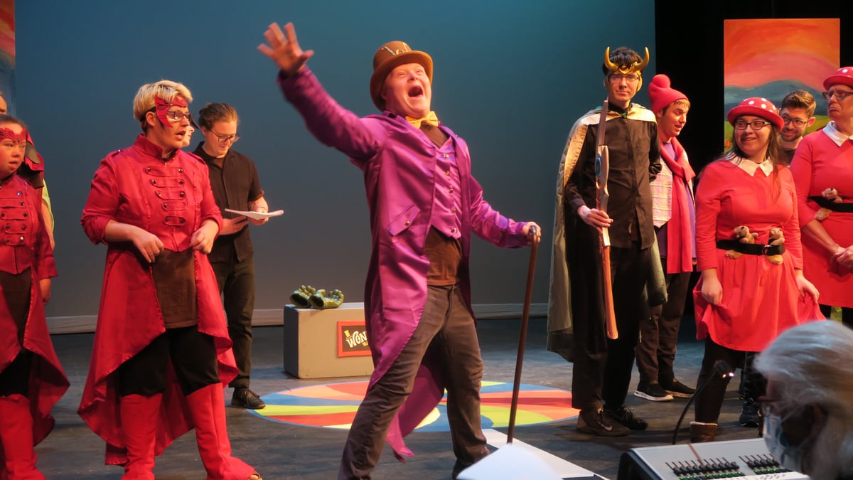 A man with Down Syndrome and a light skin tone wears a Willy Wonka costume on stage surrounded by other actors. He sings as he dramatically raises his hand upward and holds a cane in his other hand.