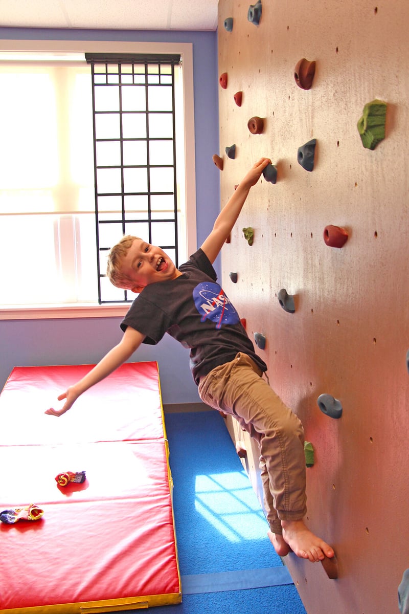 A young boy with a light skin tone hangs cheerfully with one hand from a climbing wall in the Northampton Inclusive Community Center.