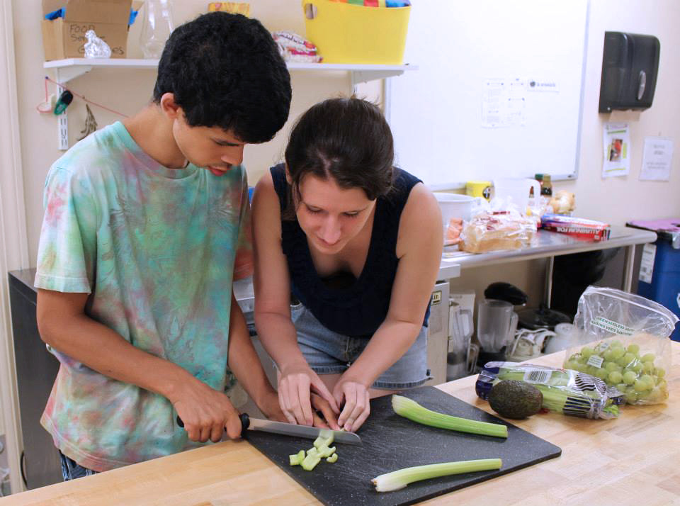 A man with a disability and a medium skin tone is cutting celery. On the right, a woman with a light skin tone is assisting him with proper finger placement.