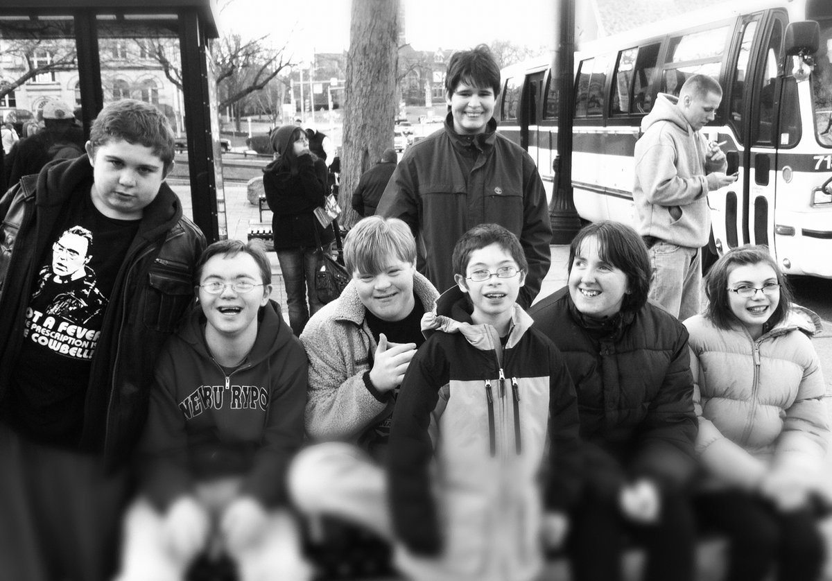 Black and white photo of a group of children with disabilities. They are standing in front of a bus station. 