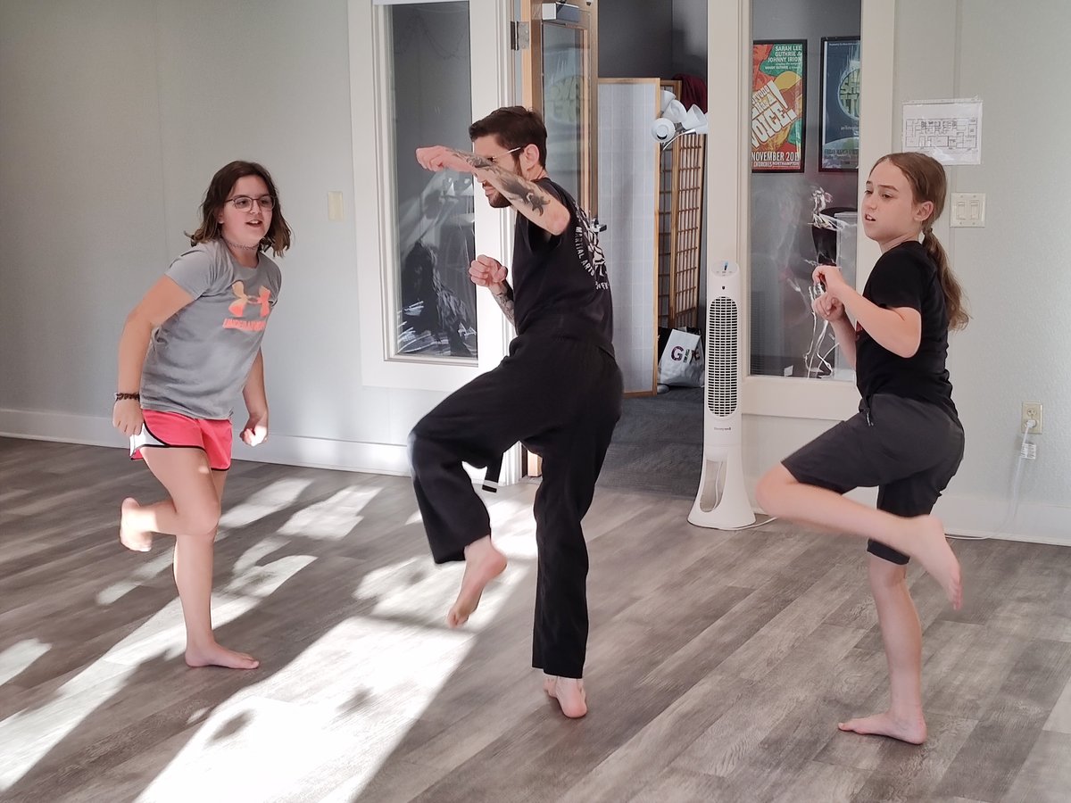 A martial arts instructor with a light skin tone stands between two students with disabilities, demonstrating a movement in class.