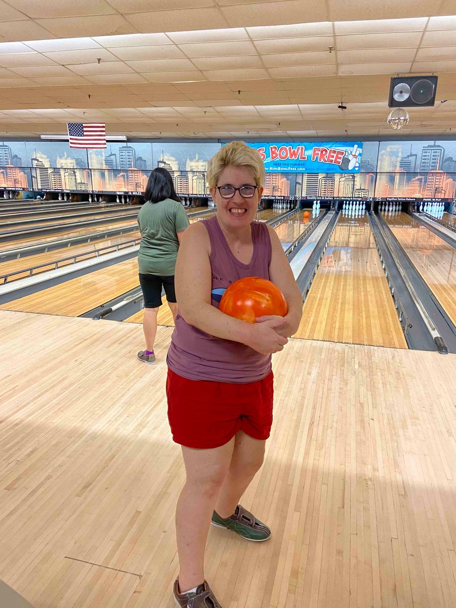 A woman with a disability and a light skin tone smiles as she holds a bowling ball in both arms.