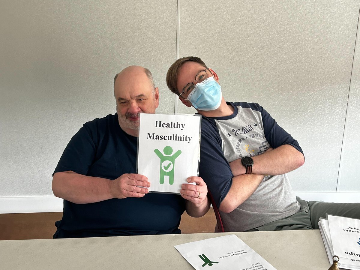 Two men with light skin tones pose for the camera while holding a sign that reads healthy masculinity.