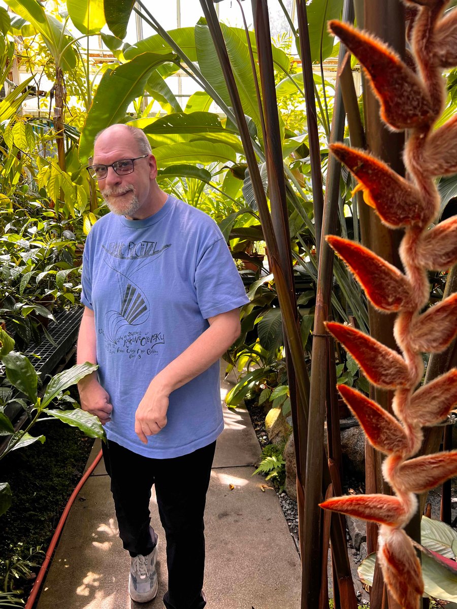 An older man with a disability and a light skin tone smiles at the camera as he walks between and underneath large plants.