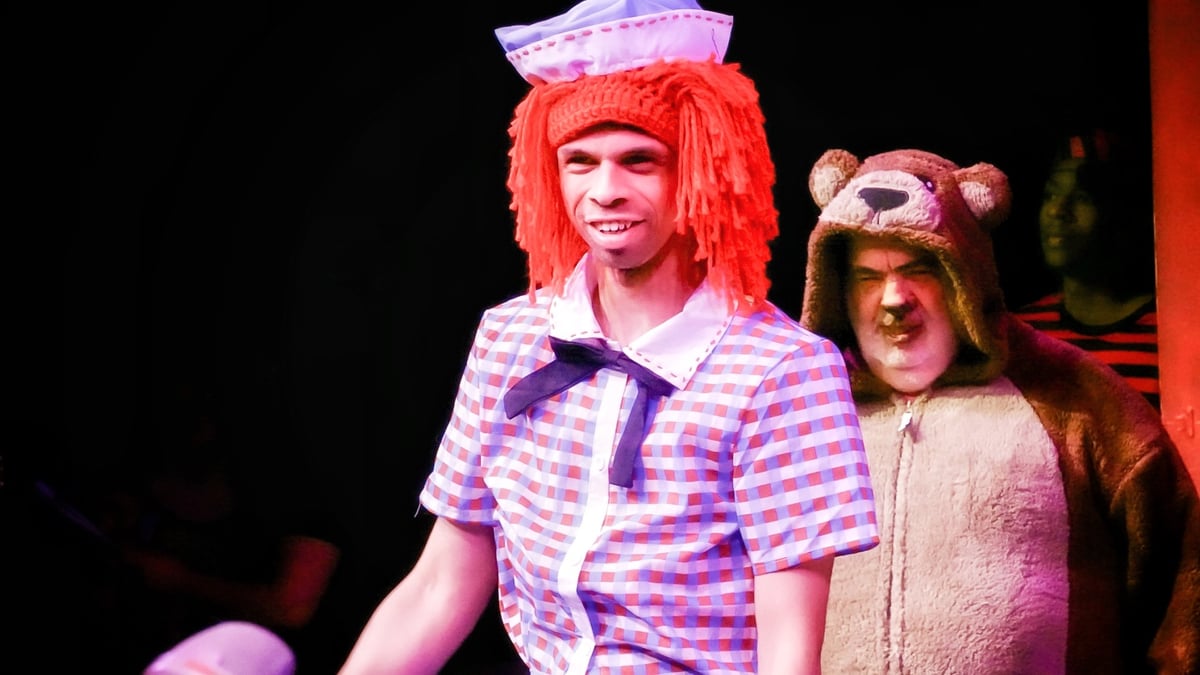 A man with a disability with a light skin tone is on a theater wearing a sailor uniform with a red, fuzzy wig. Behind him is a man dressed in a bear outfit.