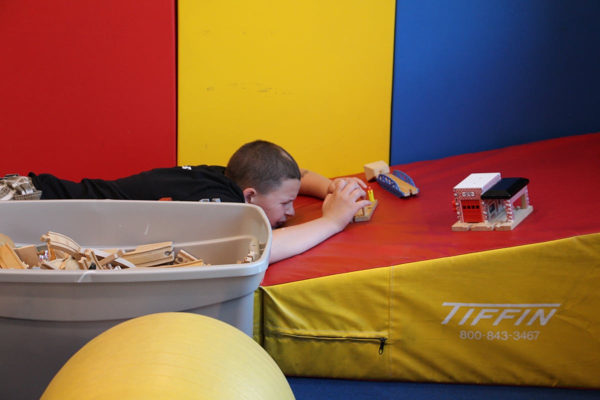 A boy with a disability with a light skin tone plays with a set of wooden train toys while laying on his stomach.